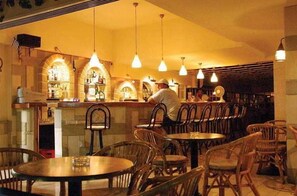 A restaurant interior with wooden tables and chairs, hanging pendant lights, and a bar area with shelves of bottles.