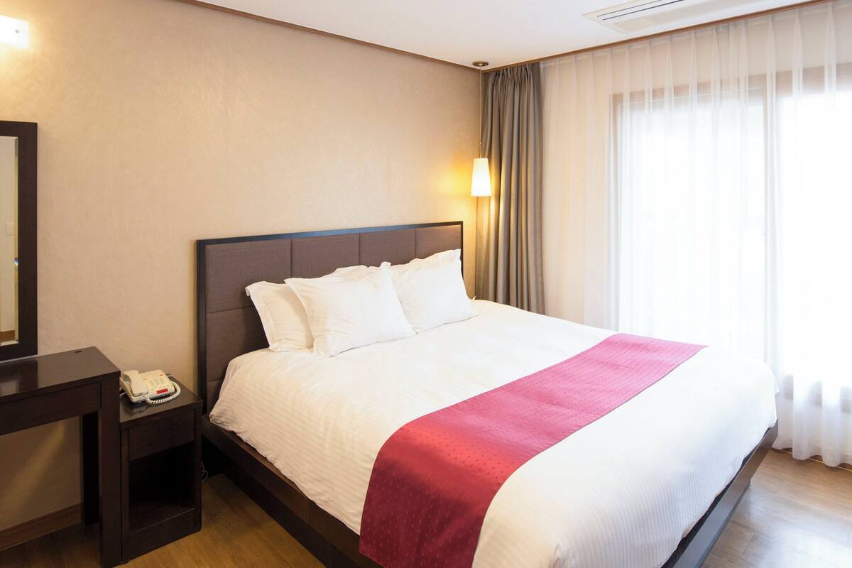 Quarto de hotel com uma cama grande, uma mesa de cabeceira com telefone e uma janela com cortinas finas.