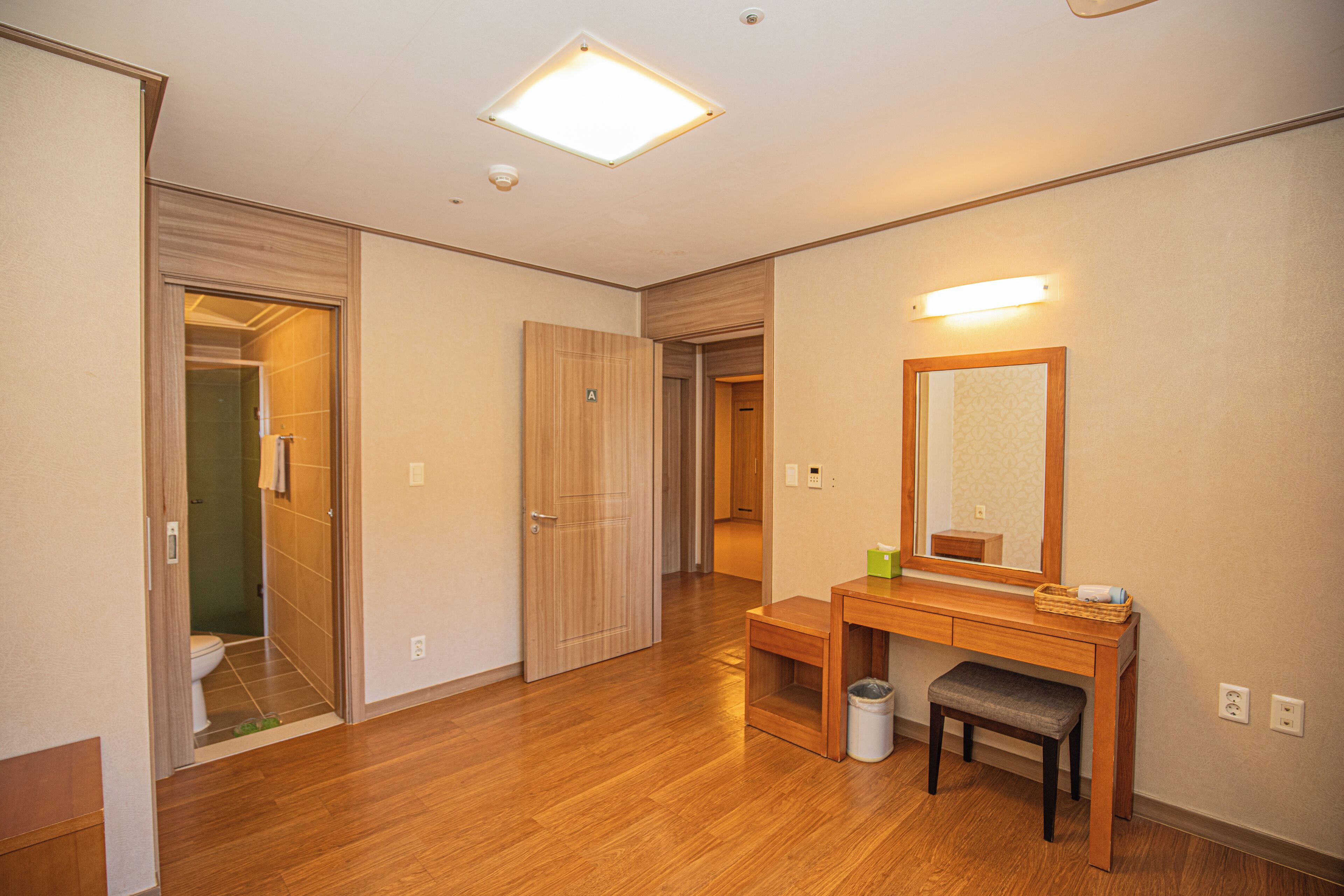A hotel room with wooden flooring, a wooden vanity with a mirror, a bench, and a trash can.