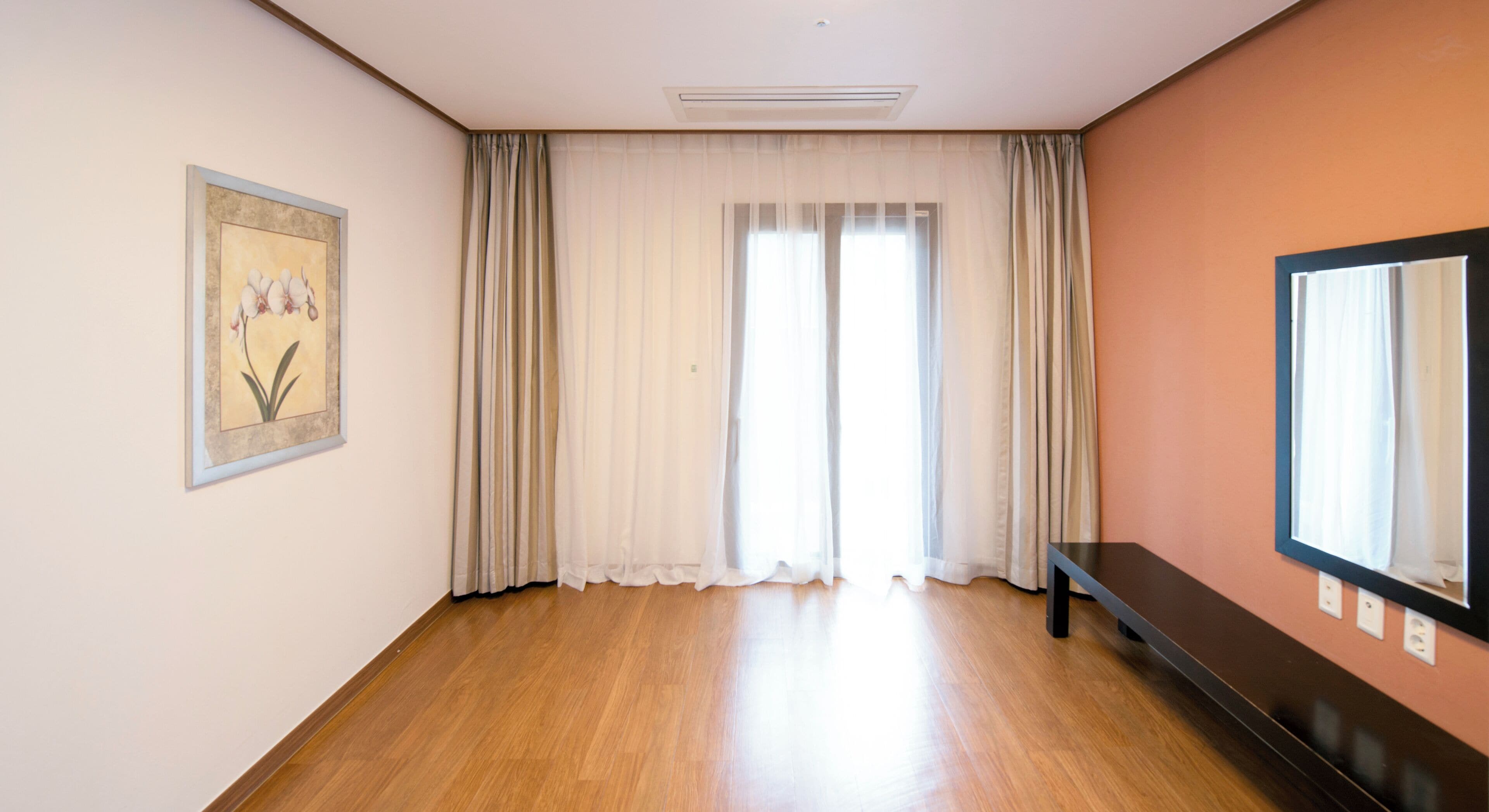 A room with wooden flooring, a large window with sheer curtains, a framed picture on the wall, and a bench along the wall.
