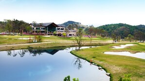 A golf course with a pond, a building with a modern design, and a hilly landscape in the background. - Holiday Inn Alpensia Pyeongchang Suites by IHG (Pyeongchang)