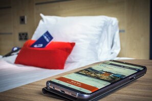 Un teléfono inteligente sobre una superficie de madera, con una aplicación de reservas de hotel abierta, un cojín rojo y una cama blanca al fondo.