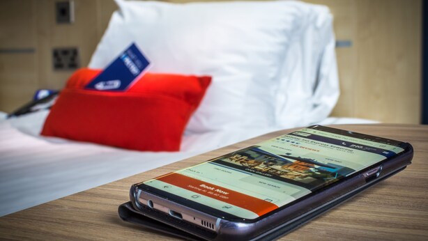 A smartphone on a wooden table with a hotel booking app open, a red pillow, and a white pillow on a bed in the background. - Holiday Inn Express Kettering by IHG (Kettering)