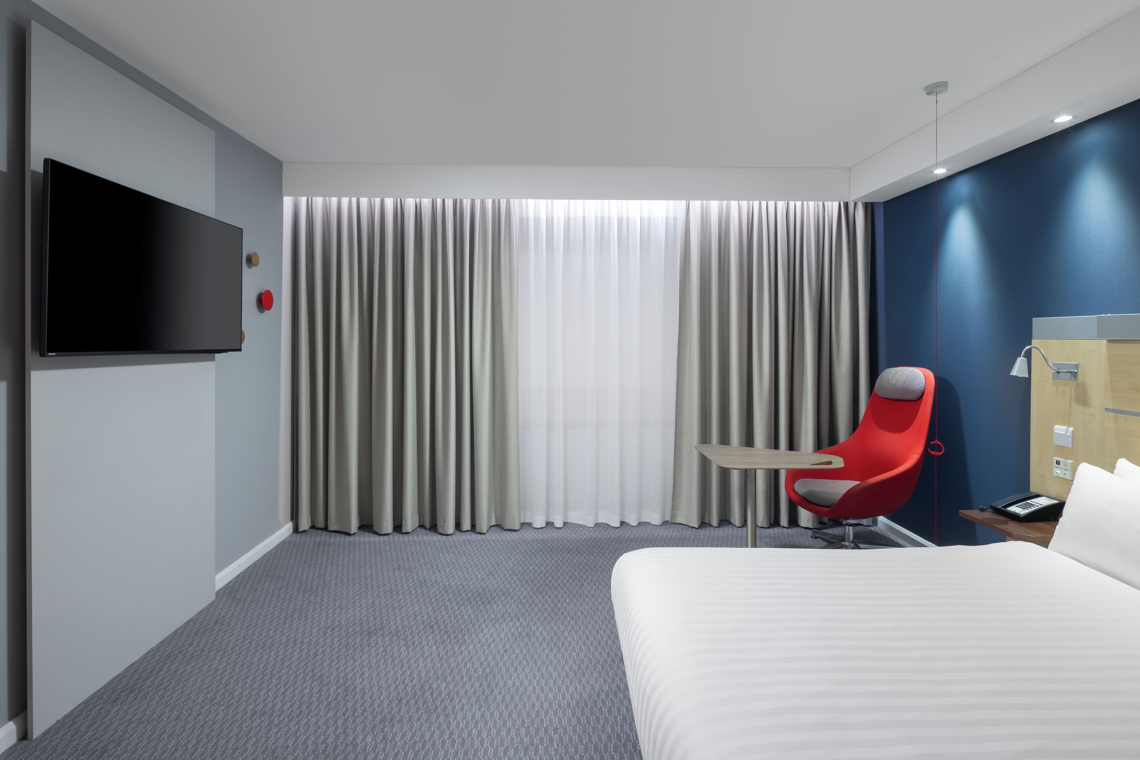 A hotel room with a bed, a red chair, a TV, and a large window with curtains.