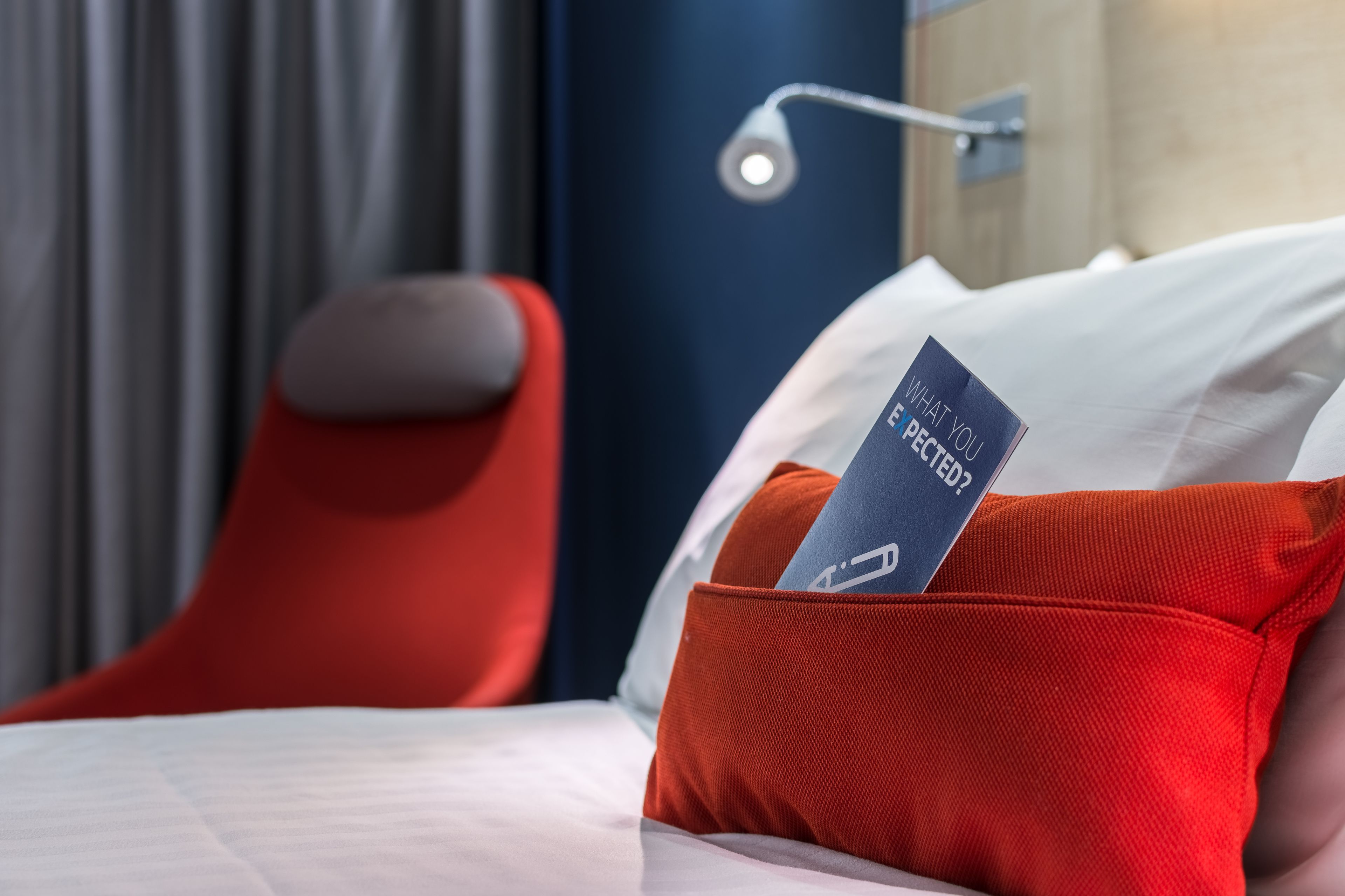 A hotel room with a bed, a red chair, and a nightstand with a lamp.