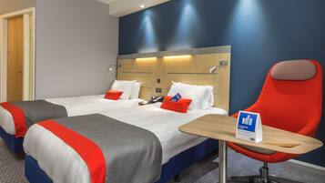 A hotel room with two beds, a red chair, a wooden table, and a sign on the table.