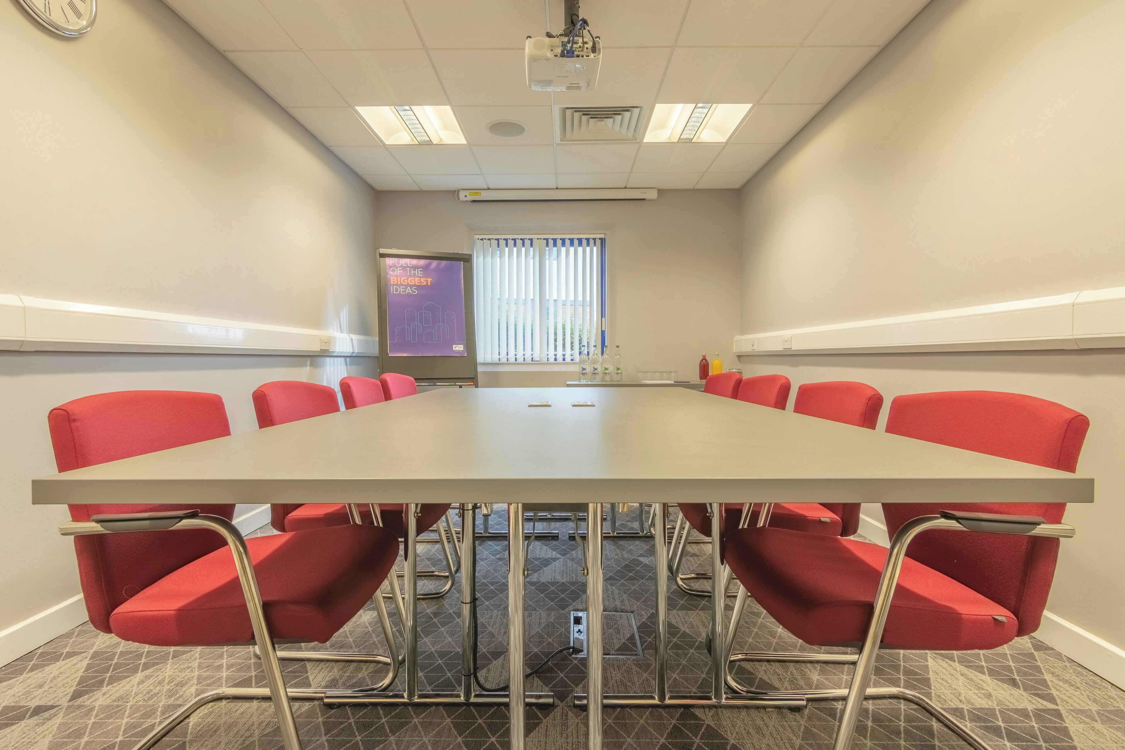 Une salle de conférence avec une longue table et des chaises rouges, un projecteur et une fenêtre équipée de stores.