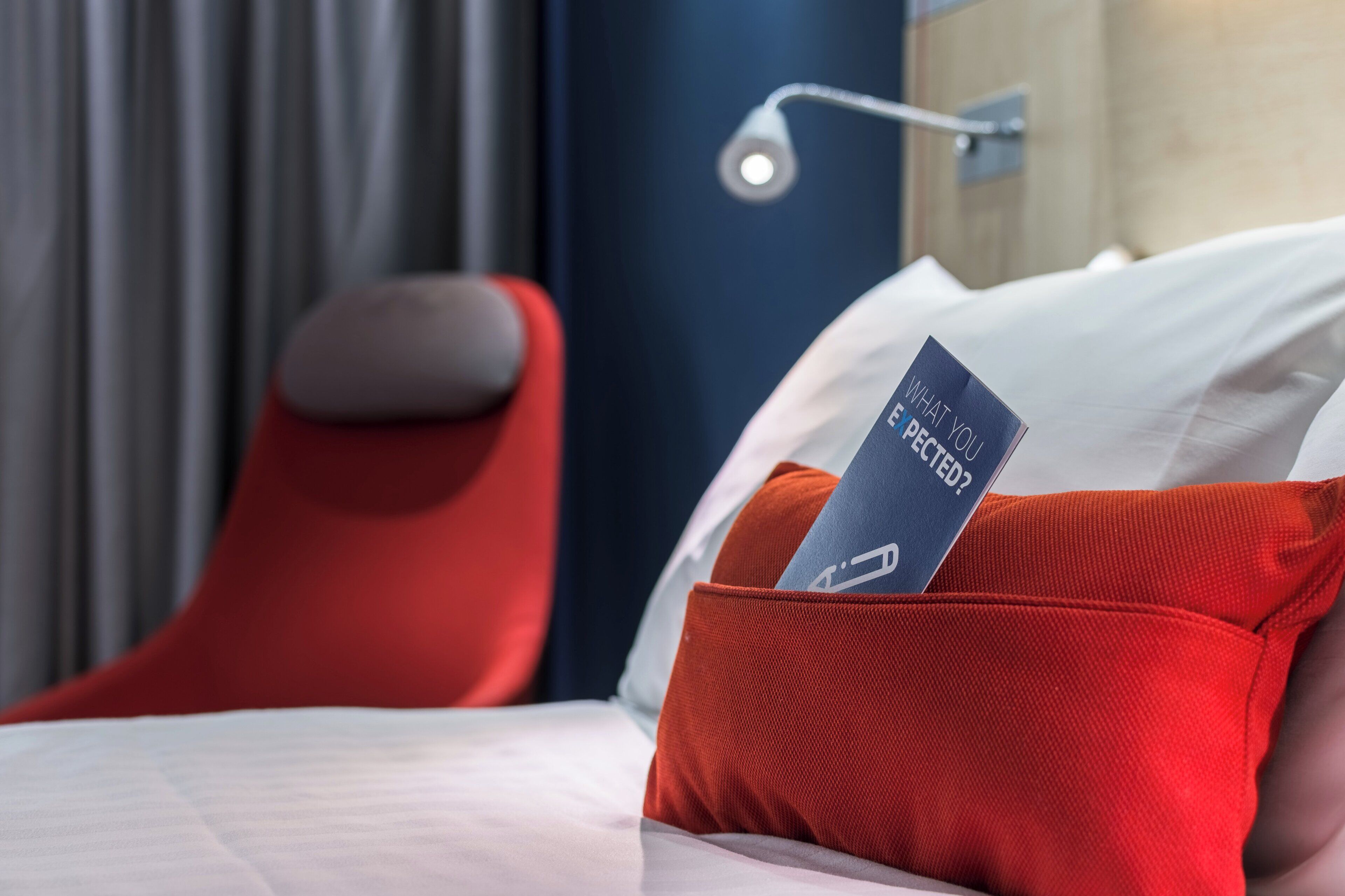 A hotel room with a bed, a red chair, a nightstand, and a wall-mounted lamp.