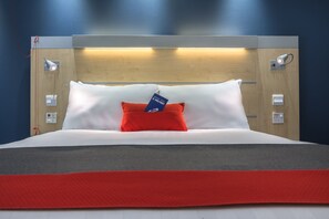 A neatly made bed with white and red bedding, a blue pillow with a tag, and a wooden headboard with integrated lighting.