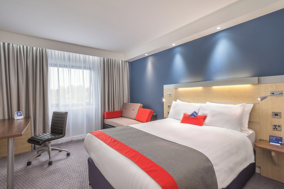 Habitación de hotel con cama, escritorio, silla y un amplio ventanal con cortinas.