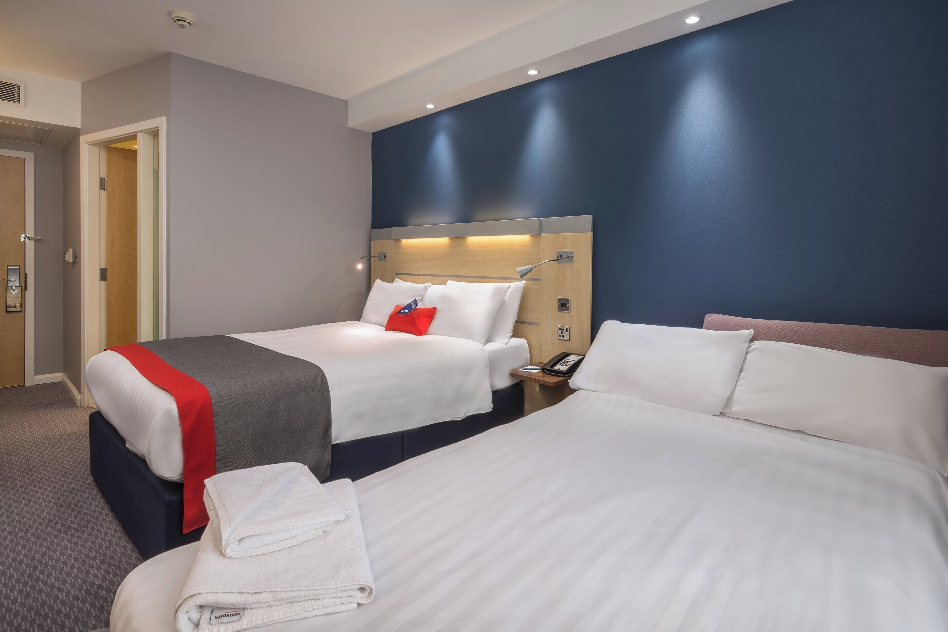 A hotel room with two beds, a red throw, a phone on the nightstand, and a door to the left.