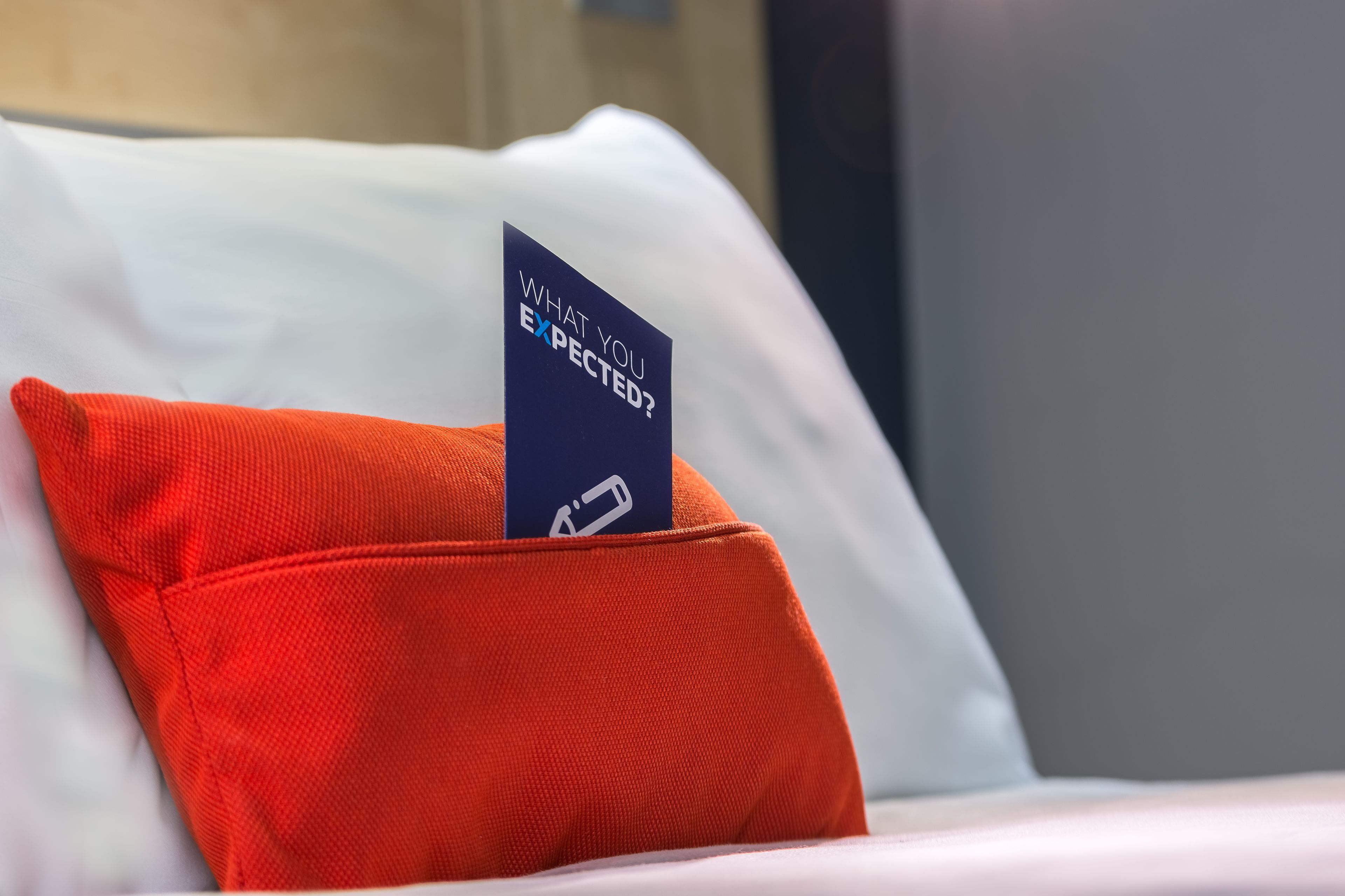 A bed with a white pillow and a red pillow, and a sign on the red pillow that reads "WHAT YOU EXPECT?".