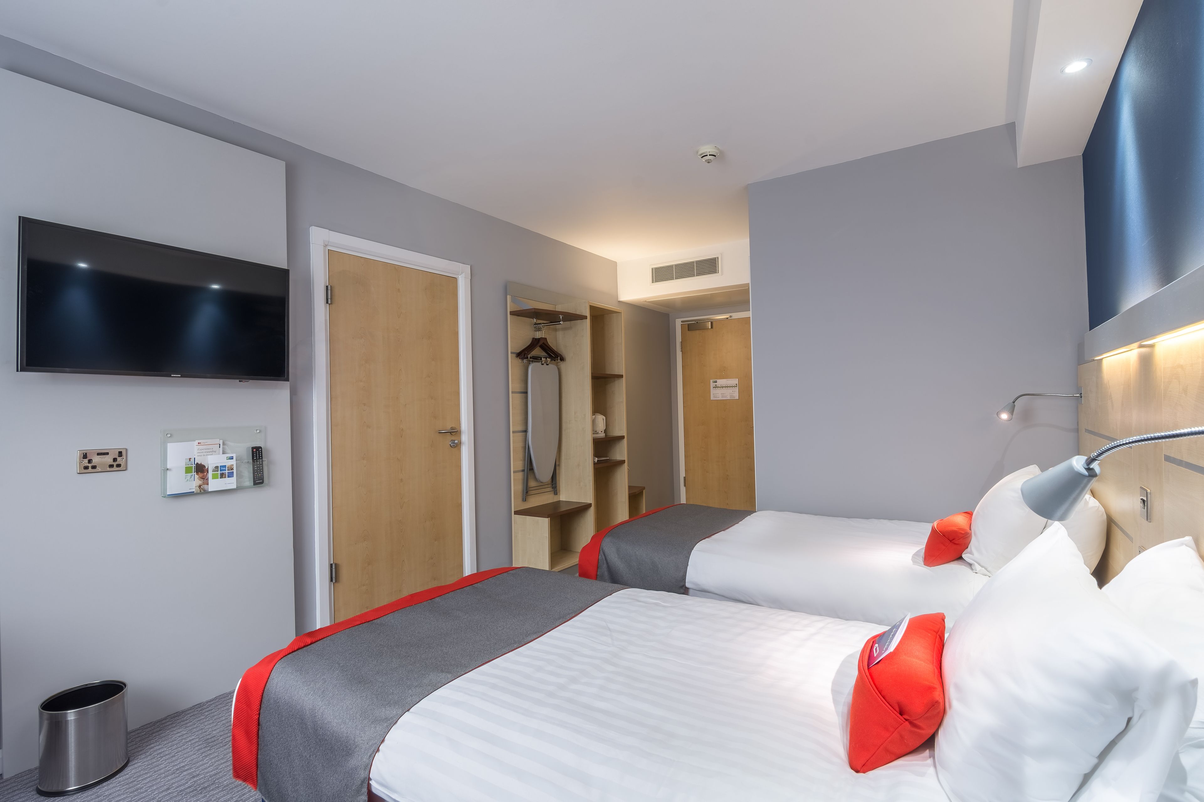 A hotel room with two beds, a TV mounted on the wall, a mirror, and a shower area.