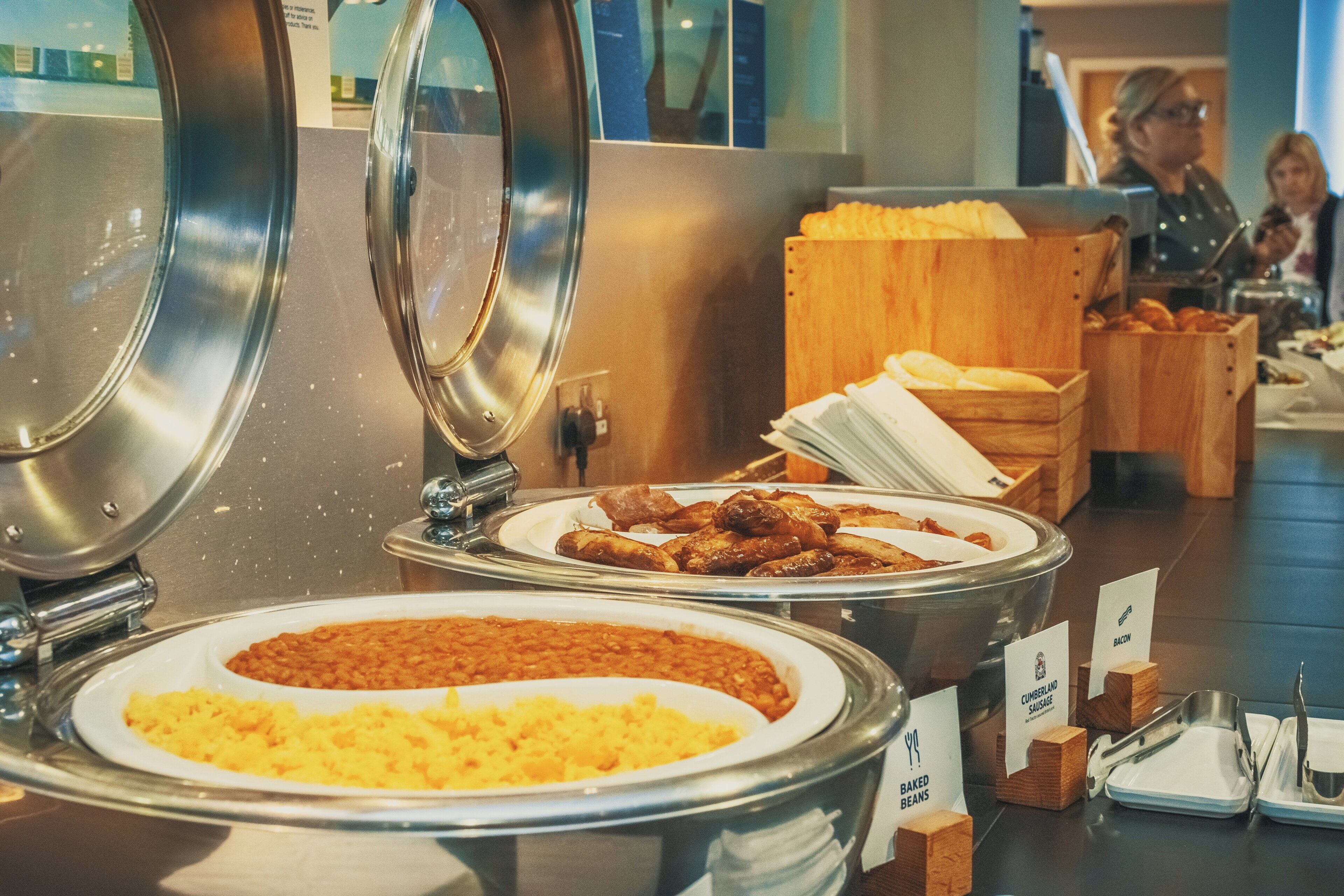 Un bufé con diversas opciones de comida, incluyendo frijoles horneados, salchicha Cumberland y otros platos.