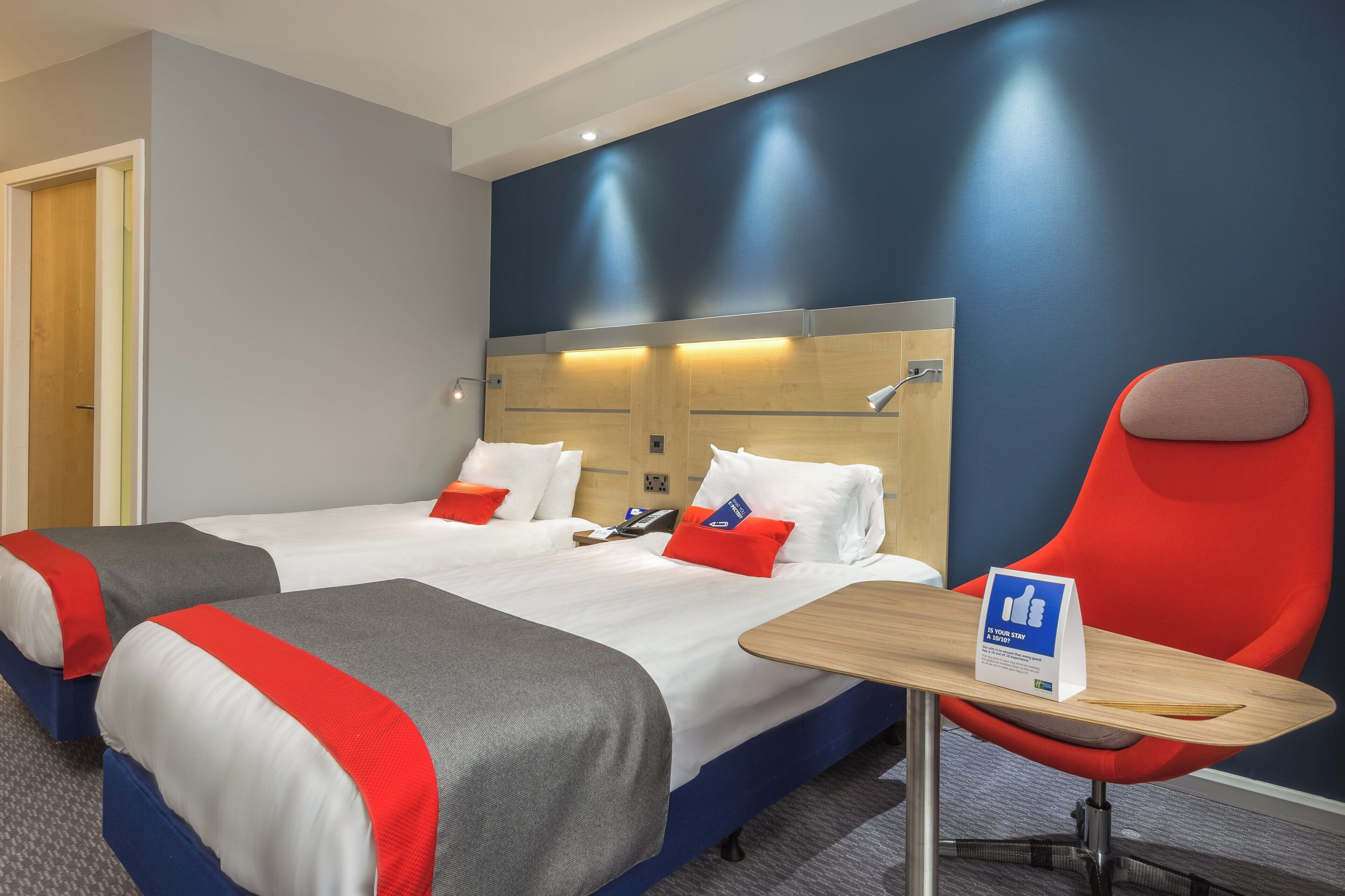 A hotel room with two beds, a red chair, a wooden table, and a sign on the table.