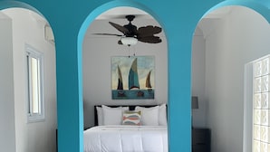 Une chambre à coucher avec un lit, un ventilateur de plafond, un tableau représentant des voiliers et une fenêtre.