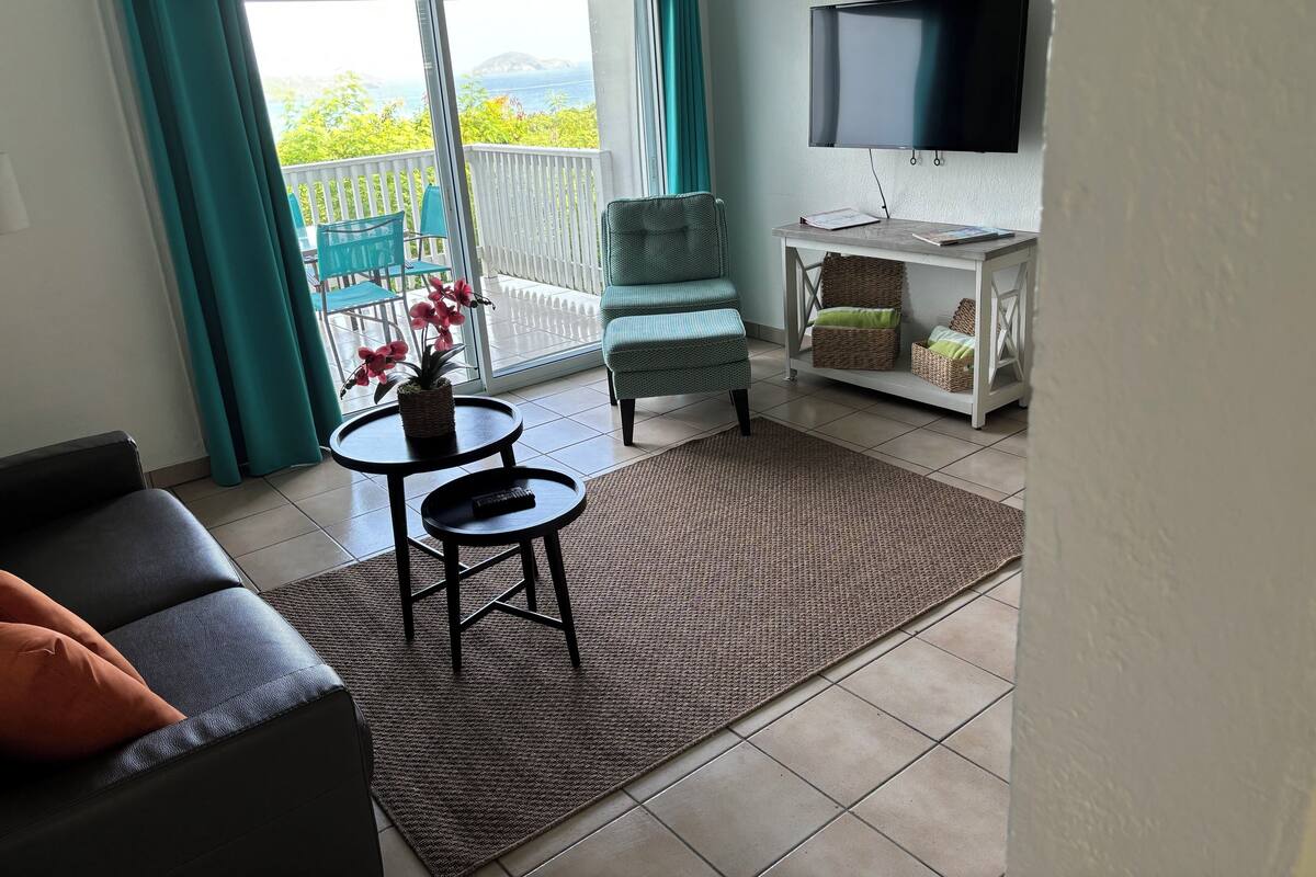 Un salon comprenant un canapé, un fauteuil, une petite table, un téléviseur et un balcon avec vue.