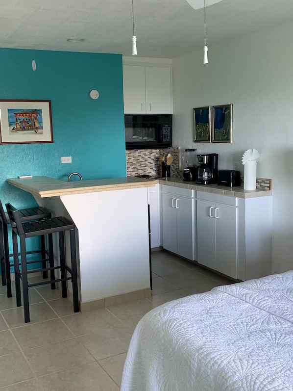 Cocina compacta con isla central, armarios blancos, microondas y cafetera. Hay dos taburetes de bar y una cama con colcha blanca.