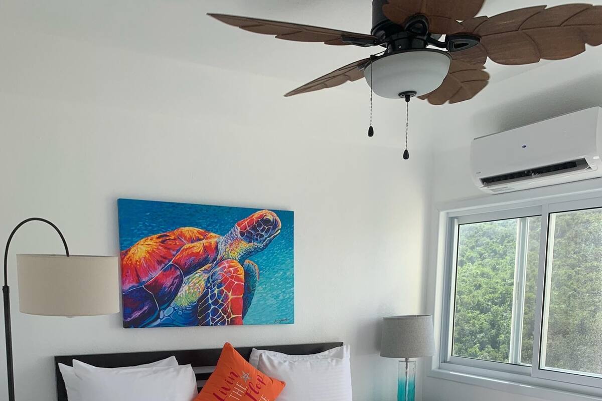 Une chambre à coucher avec un lit, un ventilateur de plafond, un tableau représentant une tortue de mer et une fenêtre donnant sur un paysage verdoyant.