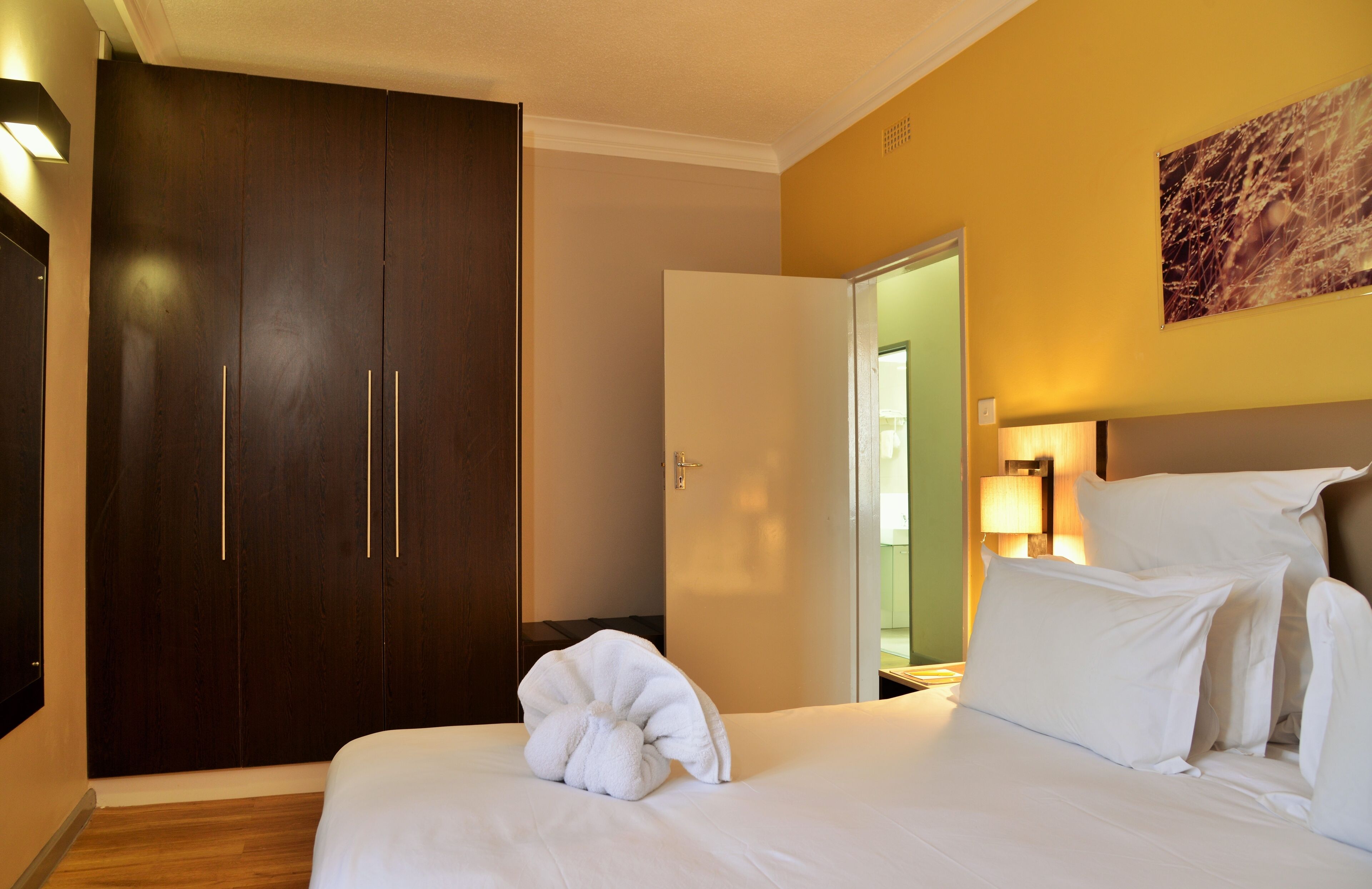 A hotel room with a bed, a wardrobe, a nightstand with a lamp, and a painting on the wall.