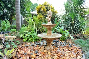 Un jardín con una fuente de piedra y una estatua, rodeado de diversas plantas y rocas.
