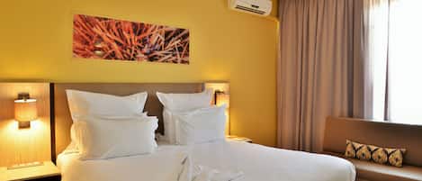 A hotel room with a large bed, two bedside lamps, a sofa with a cushion, an air conditioning unit, and a painting on the wall.