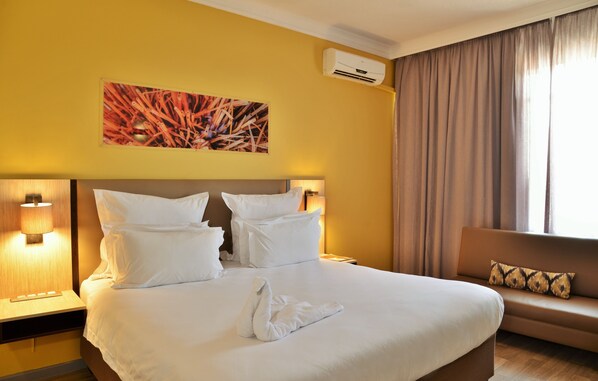 A hotel room with a large bed, two bedside lamps, a sofa with a cushion, an air conditioning unit, and a painting on the wall. - Cresta Oasis (Harare)