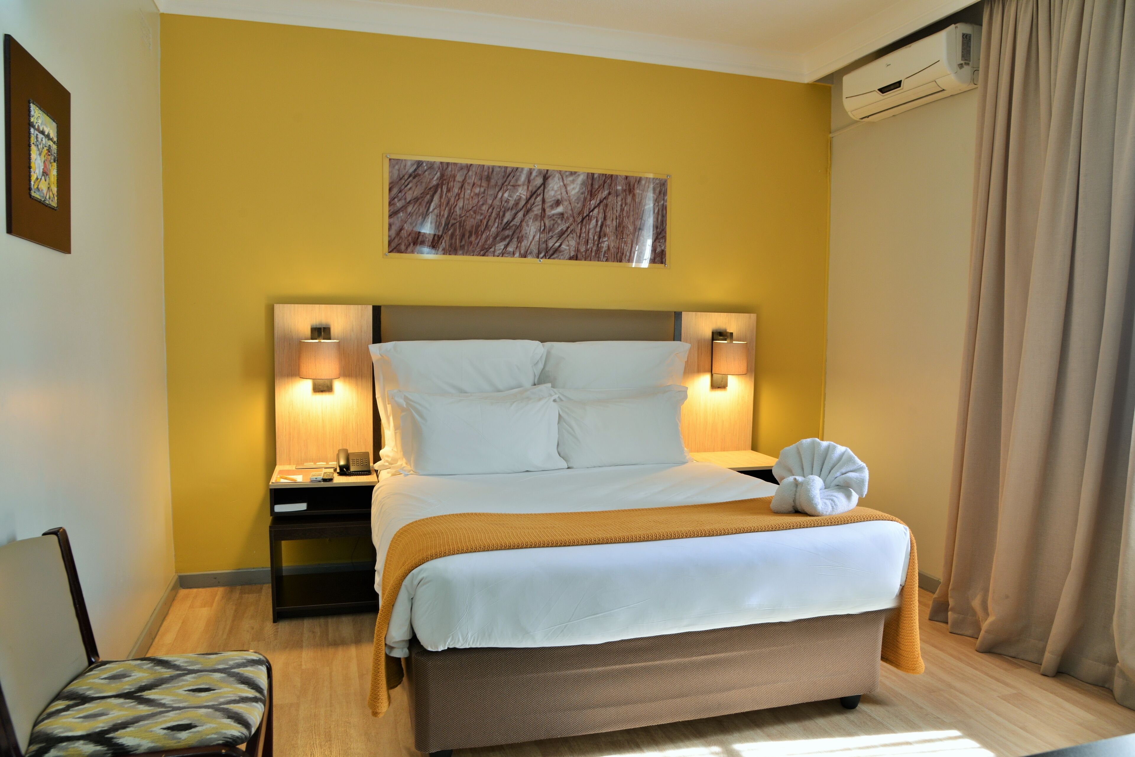 Una habitación de hotel con cama, lámparas de noche, mesita de noche, una silla y un aire acondicionado de pared.