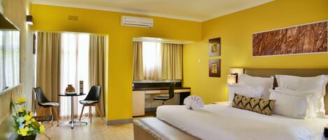 A hotel room with a large bed, a desk, and a chair. There is a window with curtains, a vase of flowers, and a painting on the wall.