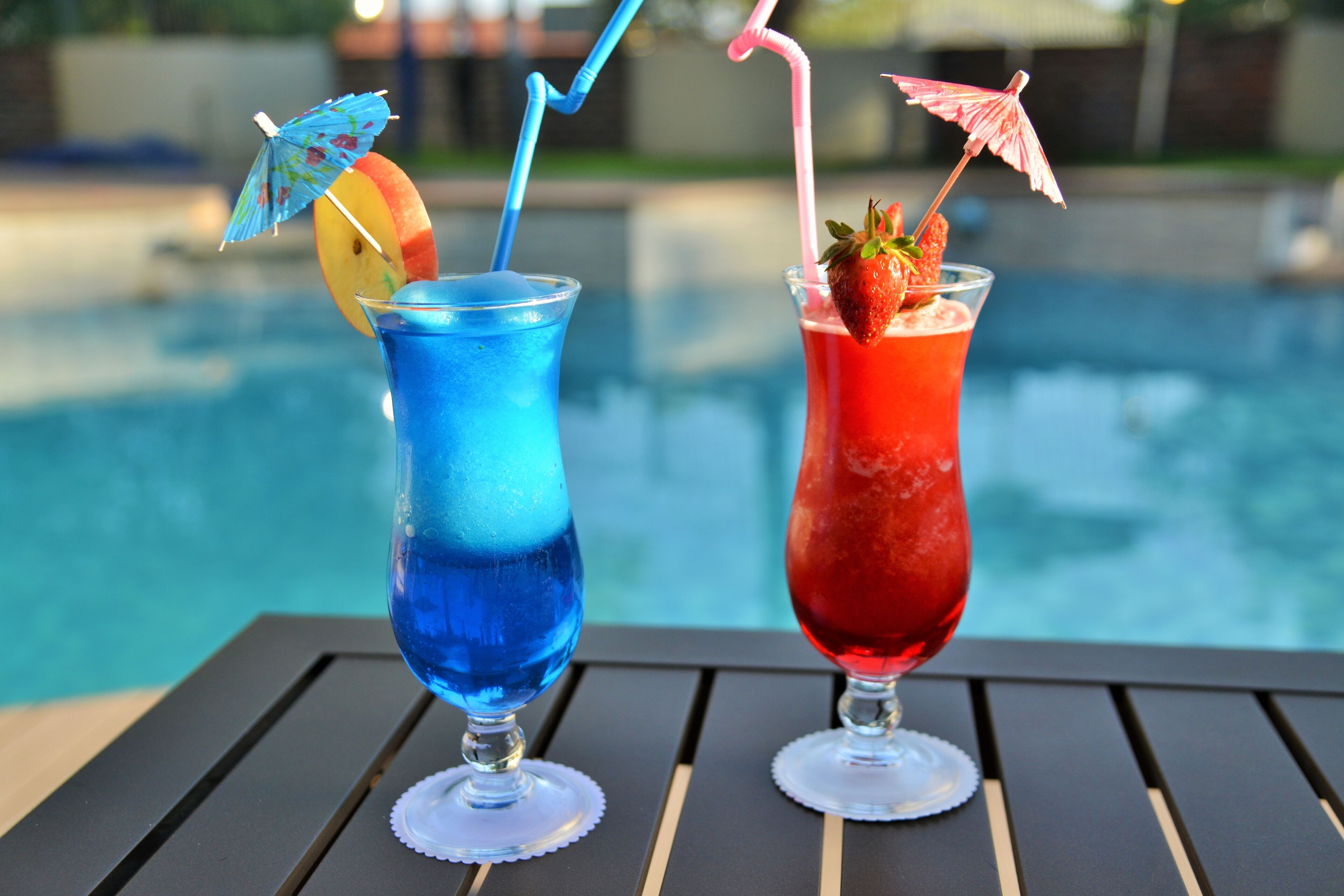Dos cócteles con pajitas y adornos de frutas sobre una mesa junto a una piscina.