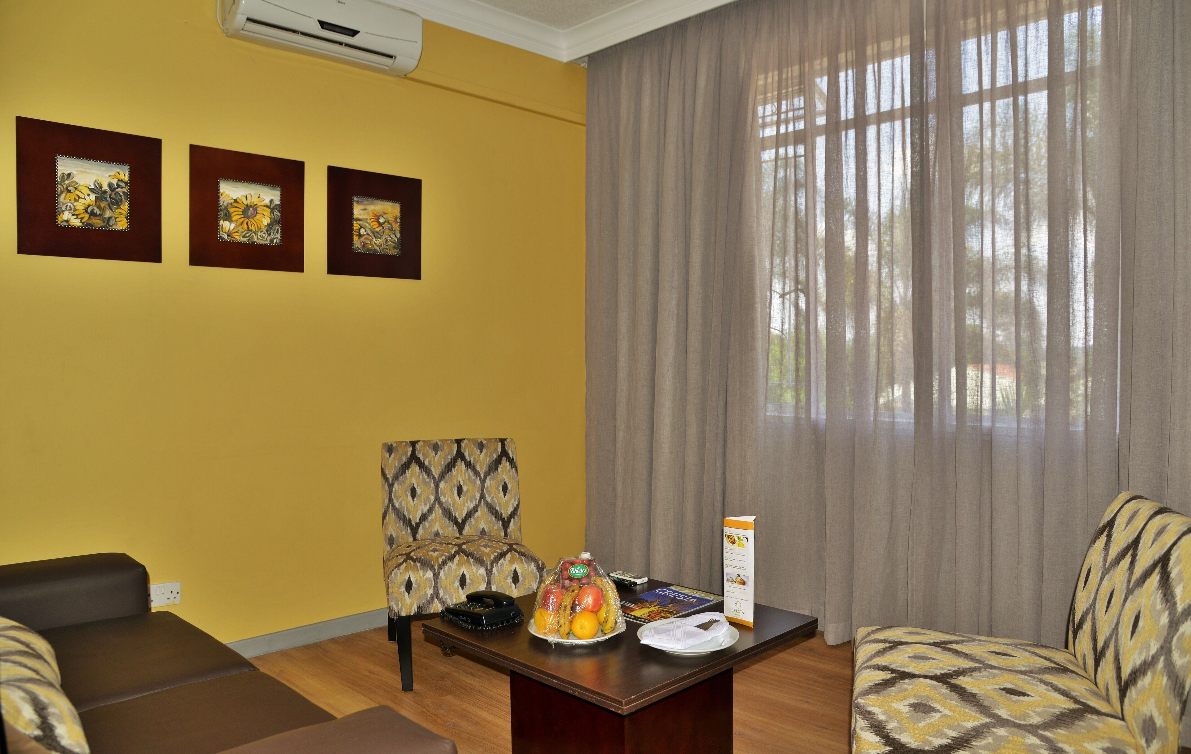 Habitación de hotel con pared amarilla, tres obras de arte enmarcadas, un sillón estampado, un sofá, una mesa de madera con una cesta de fruta, un control remoto y un atril para el menú.
