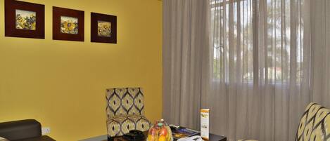 Habitación de hotel con pared amarilla, tres obras de arte enmarcadas, un sillón estampado, un sofá, una mesa de madera con una cesta de fruta, un control remoto y un atril para el menú.