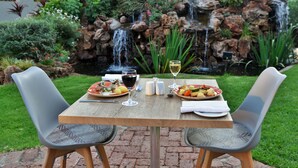 A table set outdoors with plates of food, glasses, and a bottle of wine, with a waterfall and greenery in the background.