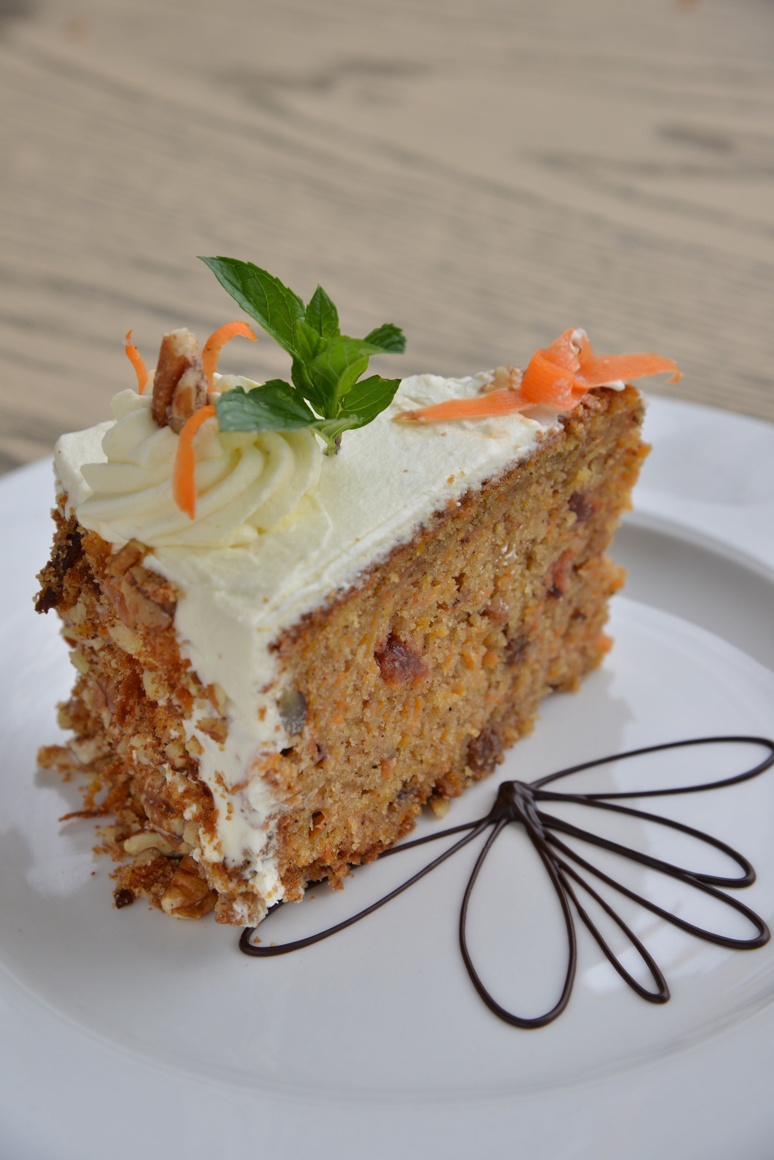 A slice of carrot cake with cream frosting and carrot garnish on a white plate.