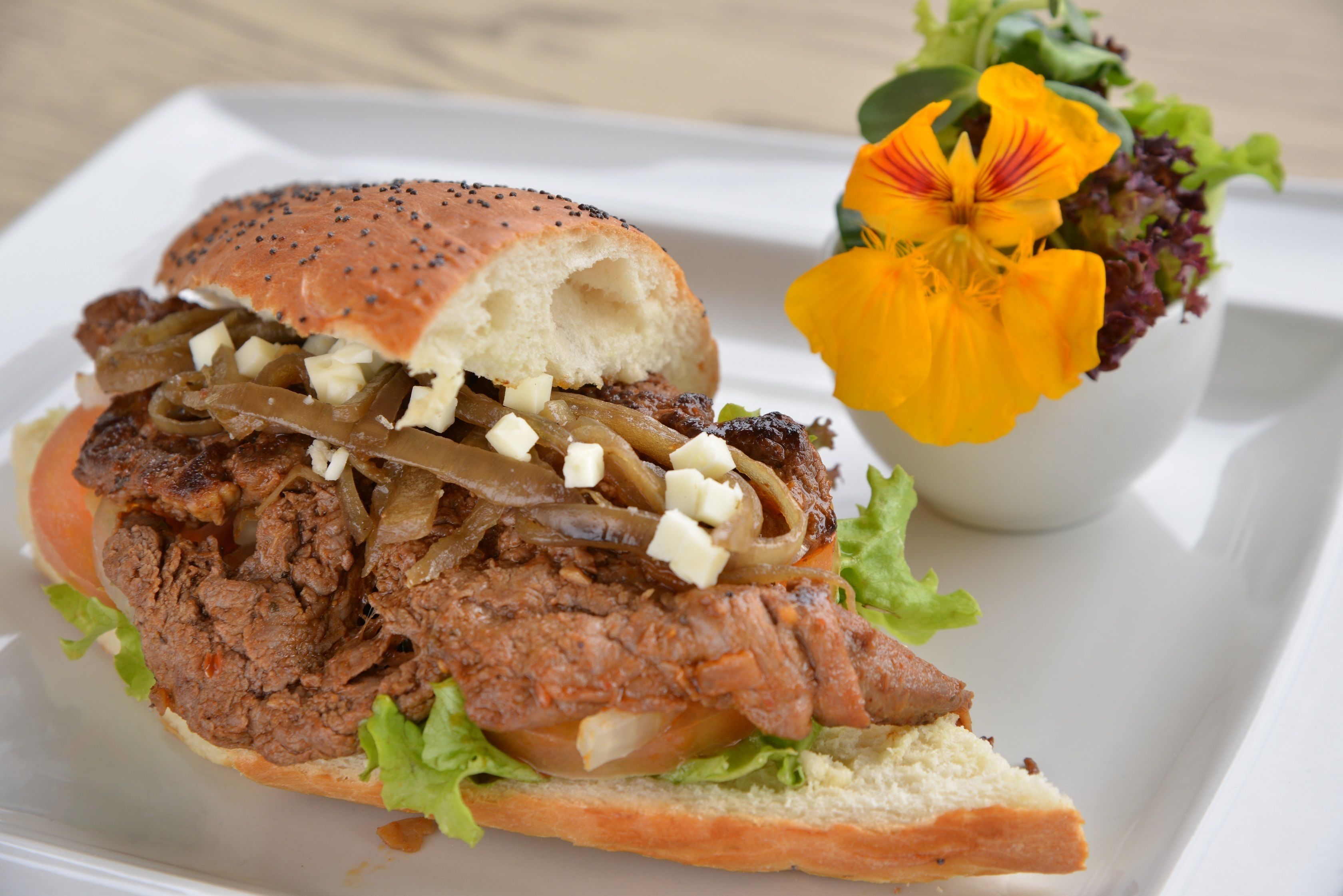 A sandwich with meat, onions, and cheese on a bun, accompanied by a flower and greens on a white plate.