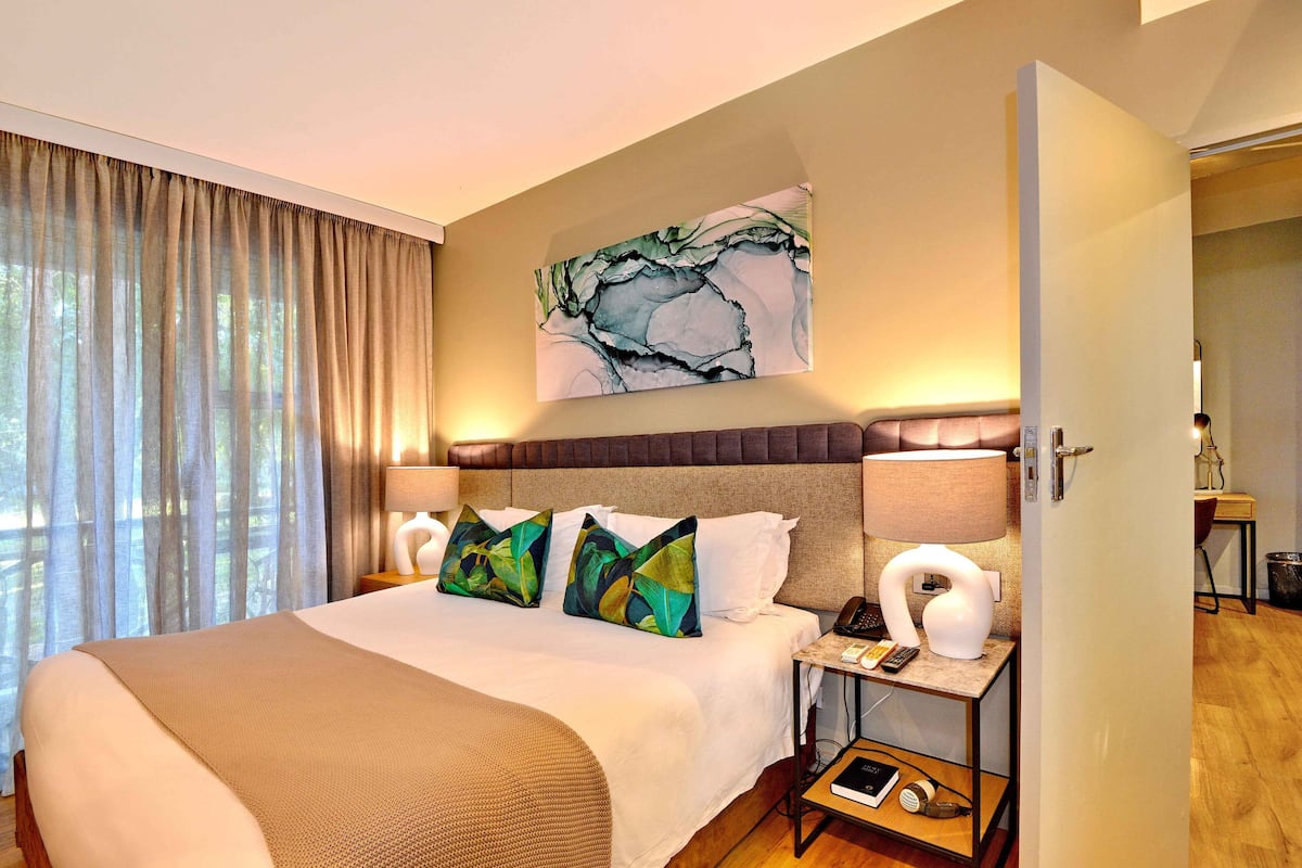 A hotel room with a large bed, a painting above it, and a nightstand with a lamp and phone.