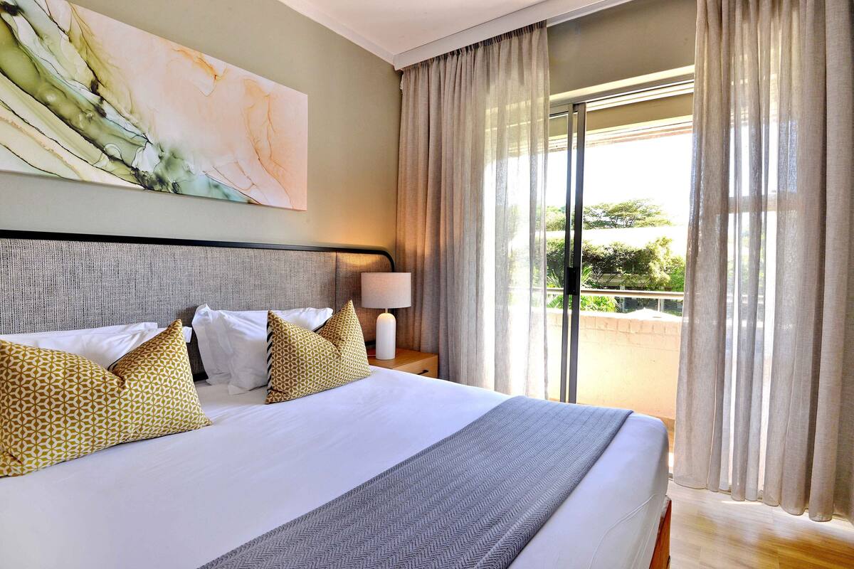 A hotel room with a large bed, a nightstand with a lamp, and a view of the outdoors through a sliding glass door.