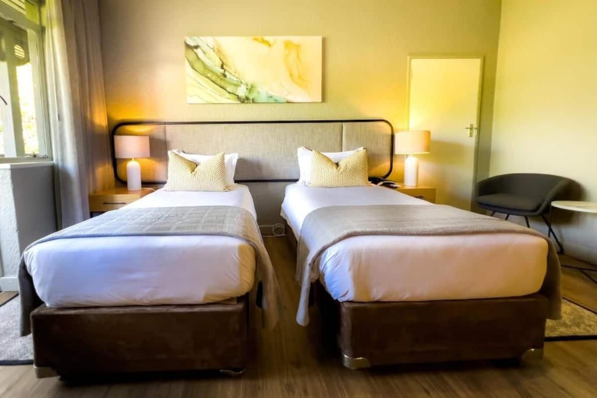 A hotel room with two beds, a headboard, bedside lamps, a chair, and a painting on the wall.