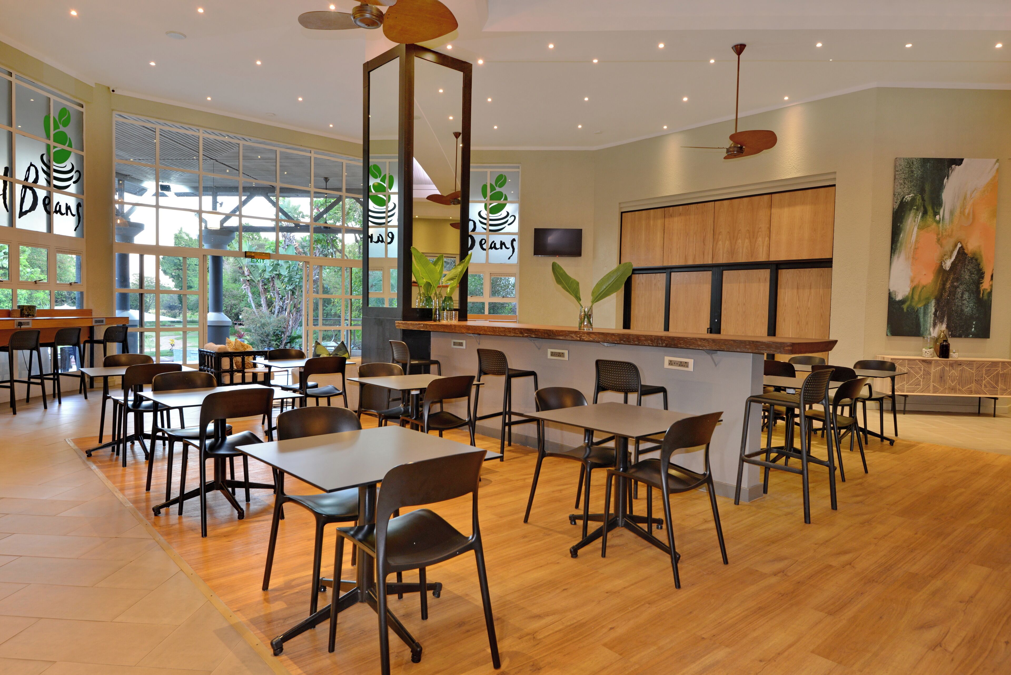 Un restaurant au décor moderne, avec des tables et des chaises en bois, de grandes fenêtres donnant sur un espace verdoyant et un comptoir équipé d’un téléviseur.