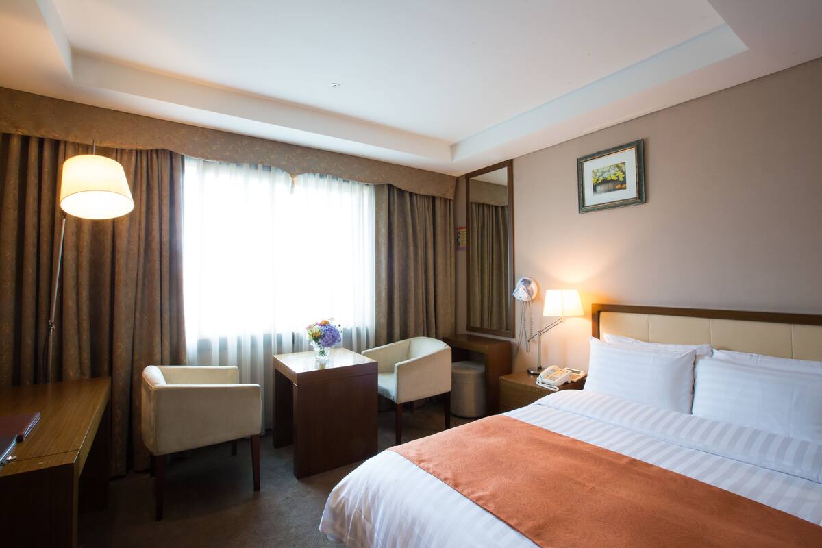 A hotel room with a bed, a desk, a chair, a lamp, and a telephone.