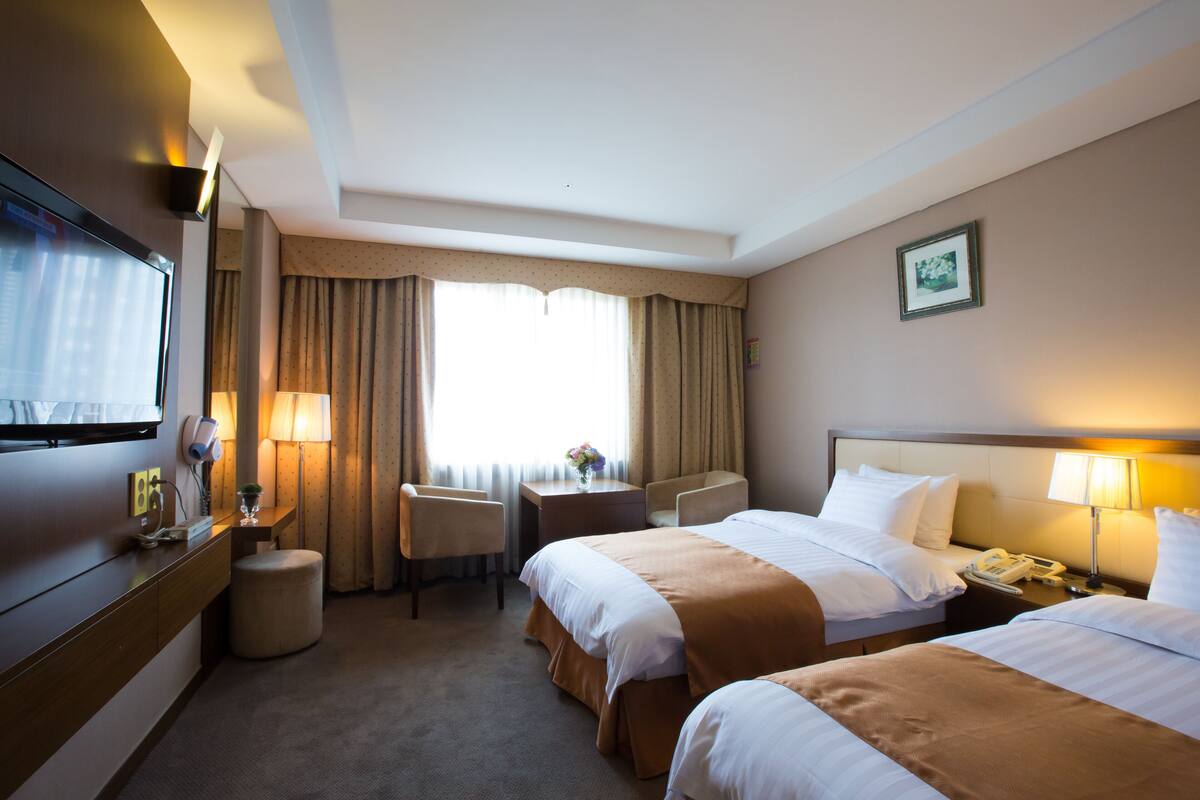 A hotel room with two beds, a desk, a chair, a TV, and a window with curtains.