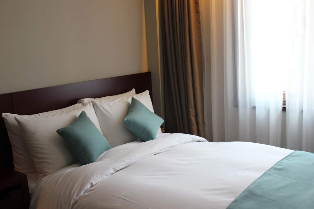 A neatly made bed with white and teal bedding, a wooden headboard, and a window with sheer curtains.