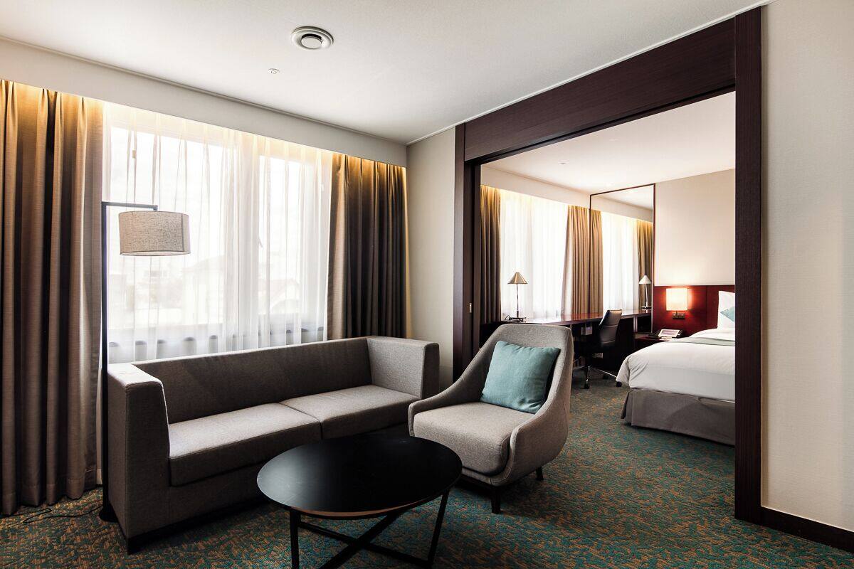 A hotel room with a sofa, armchair, and a small table. There is a bed in the background and a window with curtains.