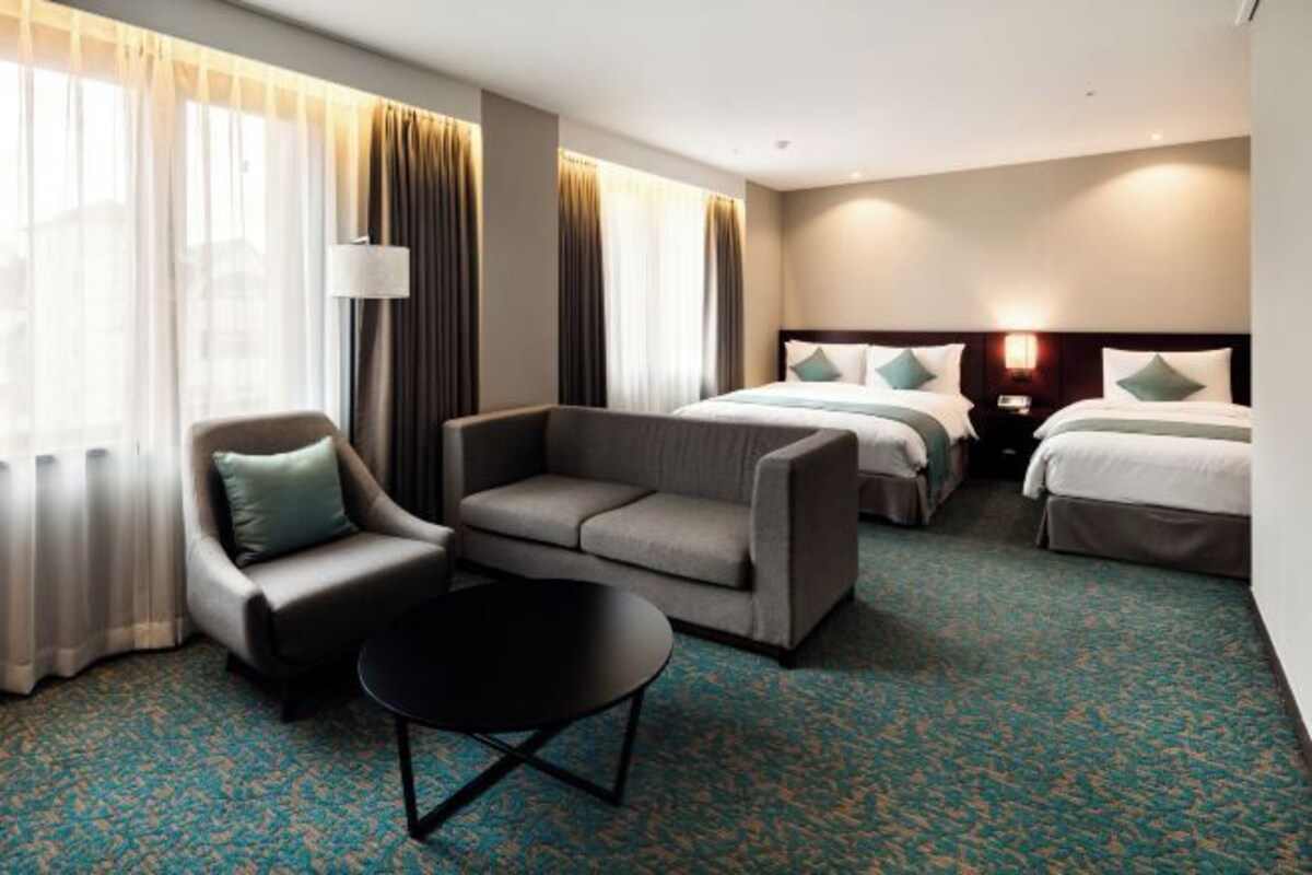 A hotel room with a sofa, two single beds, a small table, and a lamp.