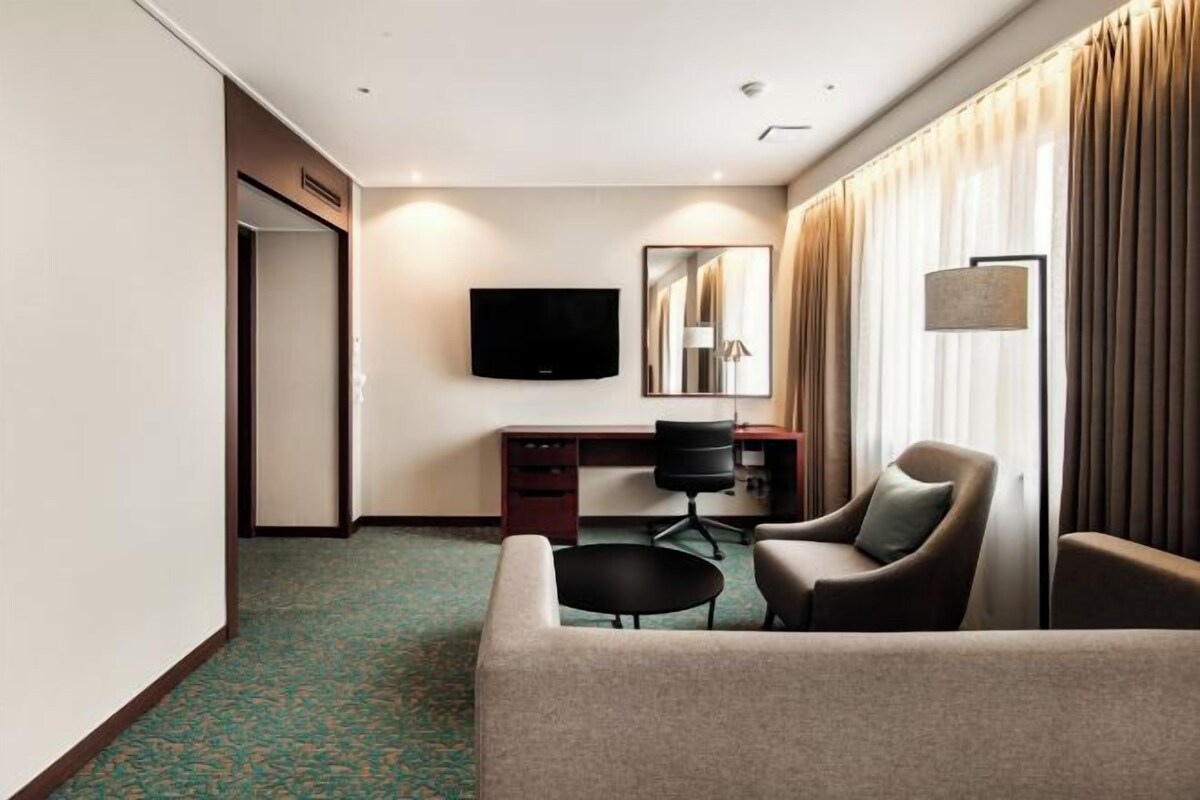Una habitación de hotel con un sofá, un escritorio con silla, una televisión y un espejo grande.