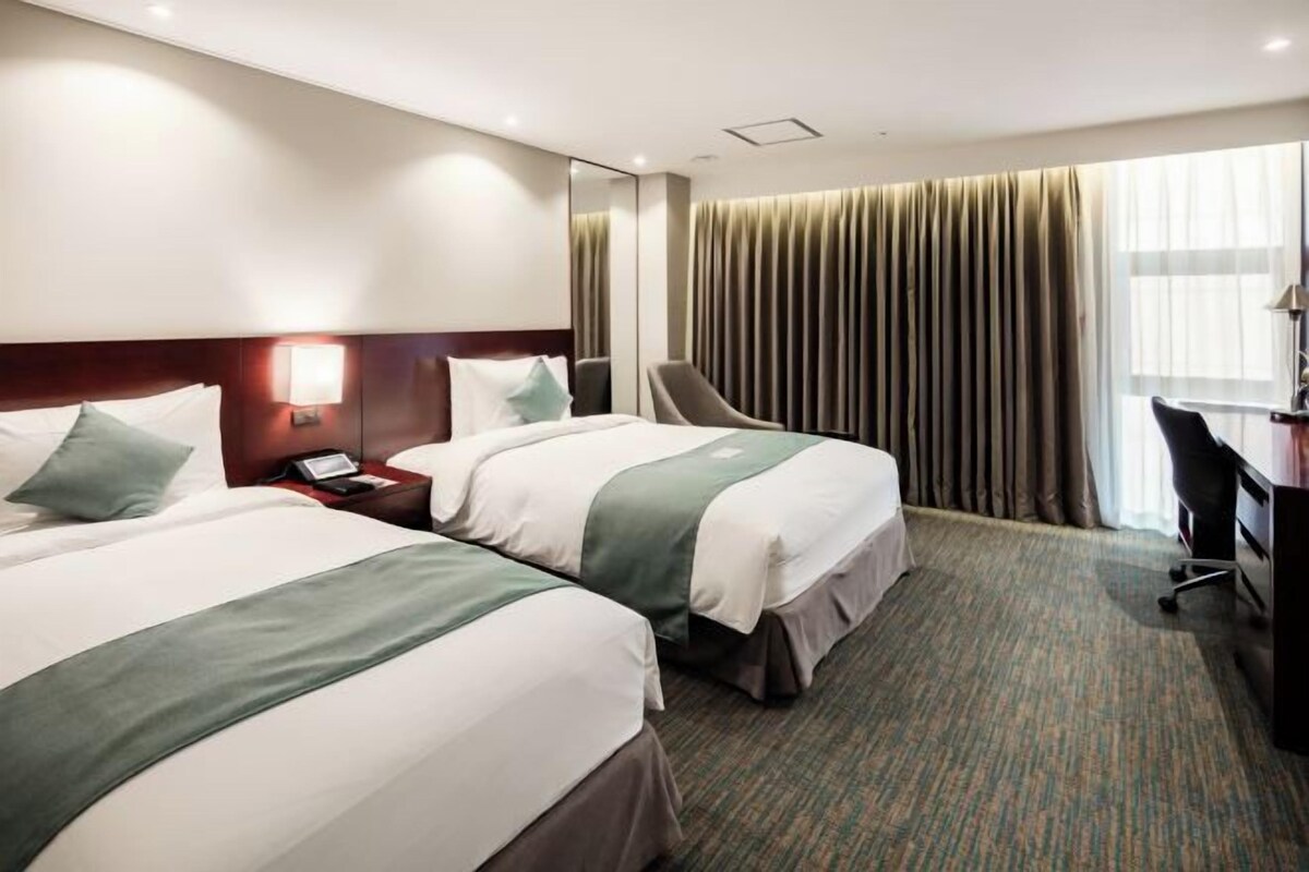 Habitación de hotel con dos camas, un escritorio, una silla y un amplio ventanal con cortinas.