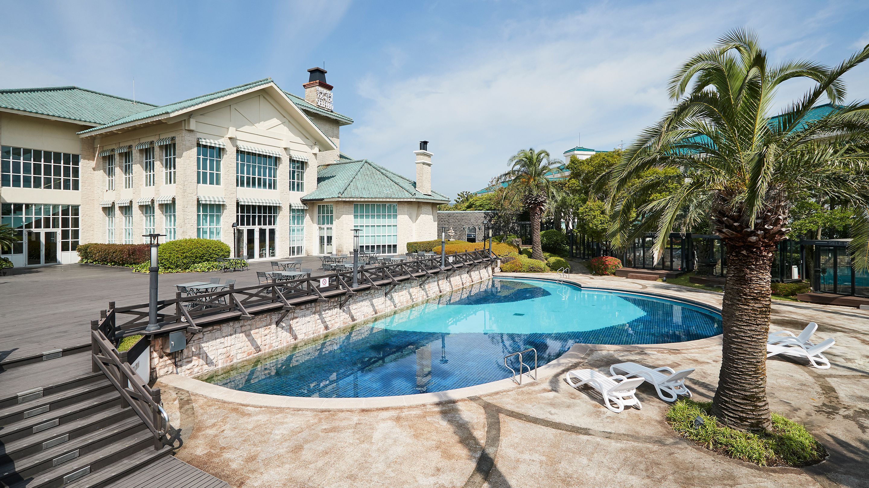 A swimming pool with lounge chairs and a wooden deck leading to a building with a green roof and stone facade.