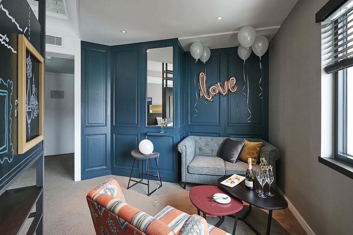 A cozy living room with a blue wall, a gray sofa, a patterned armchair, a small round table with a bottle and glasses, and a mirror reflecting a bed.