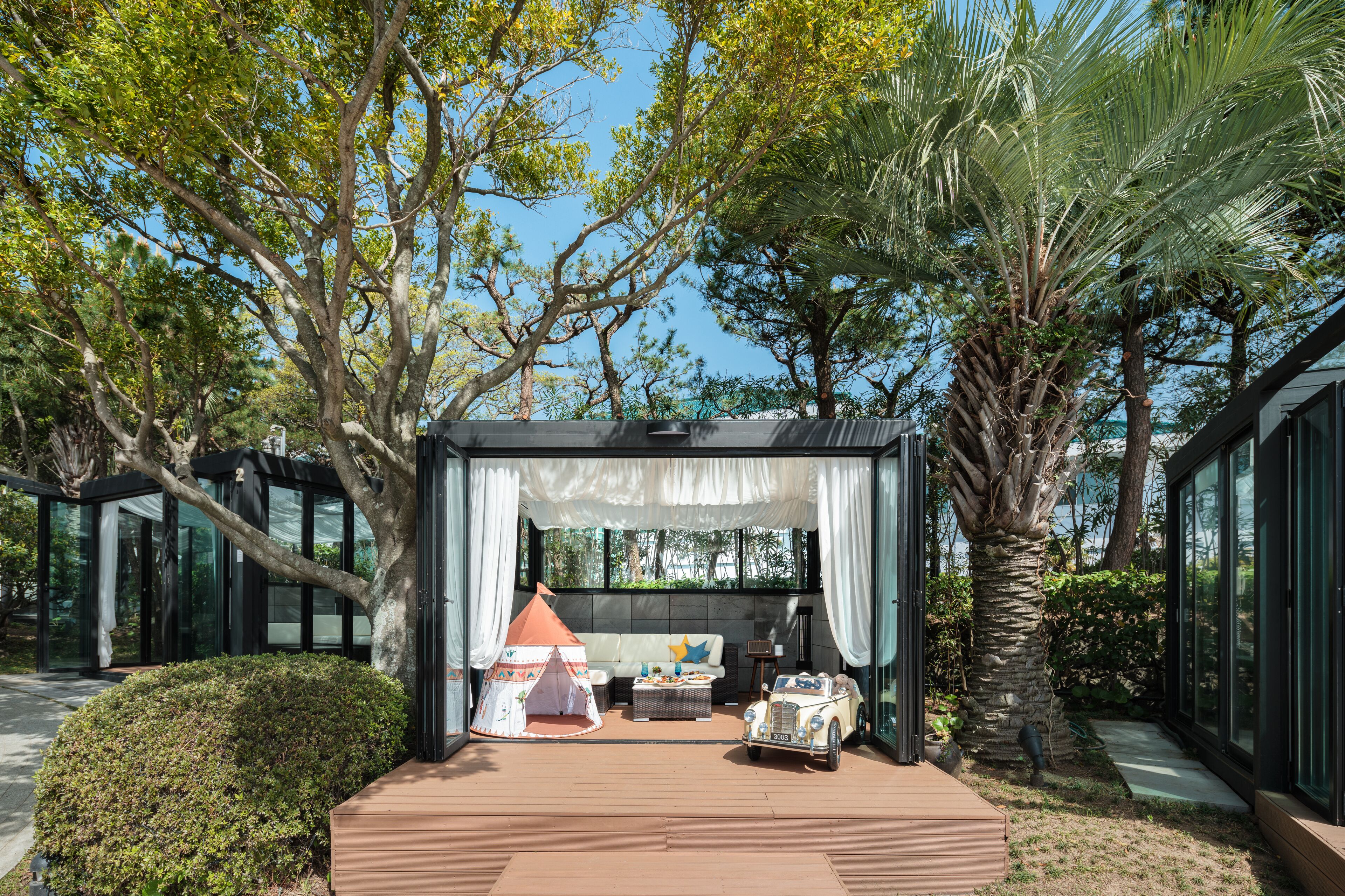 A modern outdoor living area with a wooden deck, a tent, a vintage car, and a palm tree.
