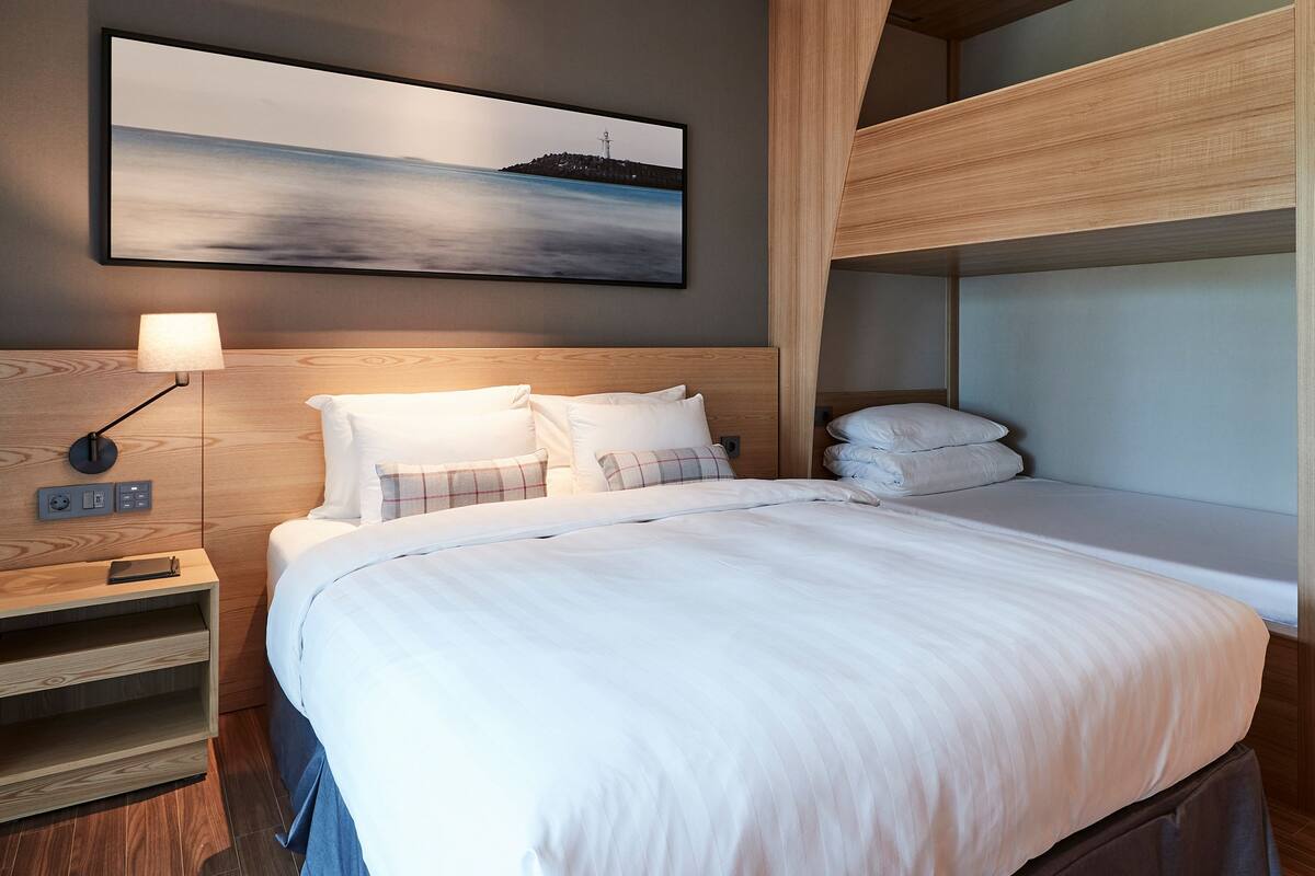 A bunk bed room with a wooden headboard, a bedside table with a lamp, and a painting of a coastal scene.